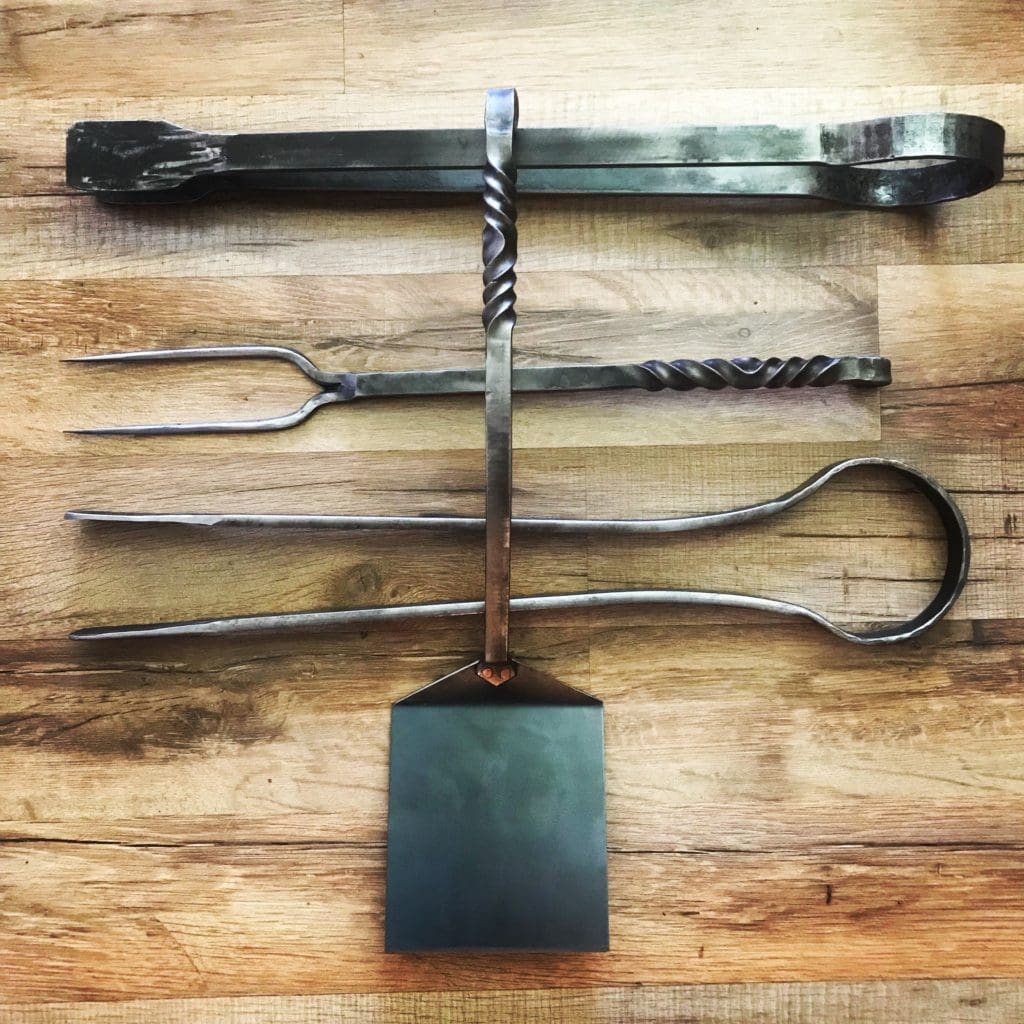 Hand forged set of grilling tools on a wood background including tongs, fork, and spatula