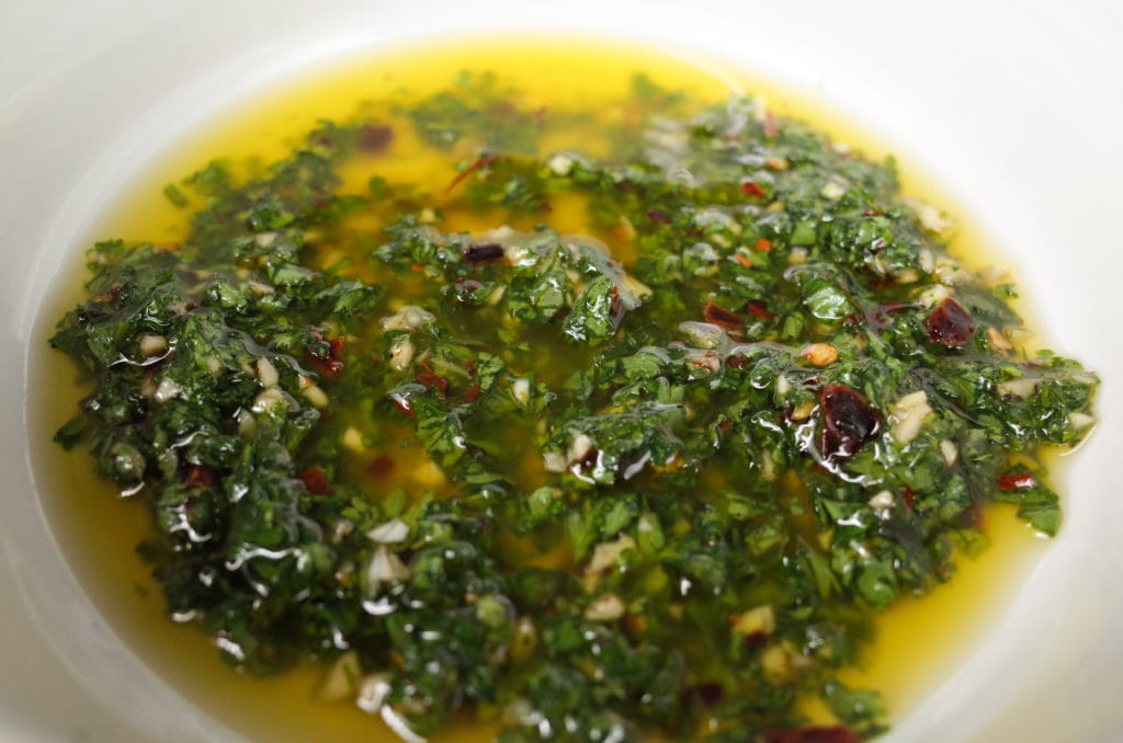 Prepared chimichurri of olive oil, chopped parsley, red pepper flakes, oregano, red wine vinegar, and minced garlic served in a white ceramic bowl.